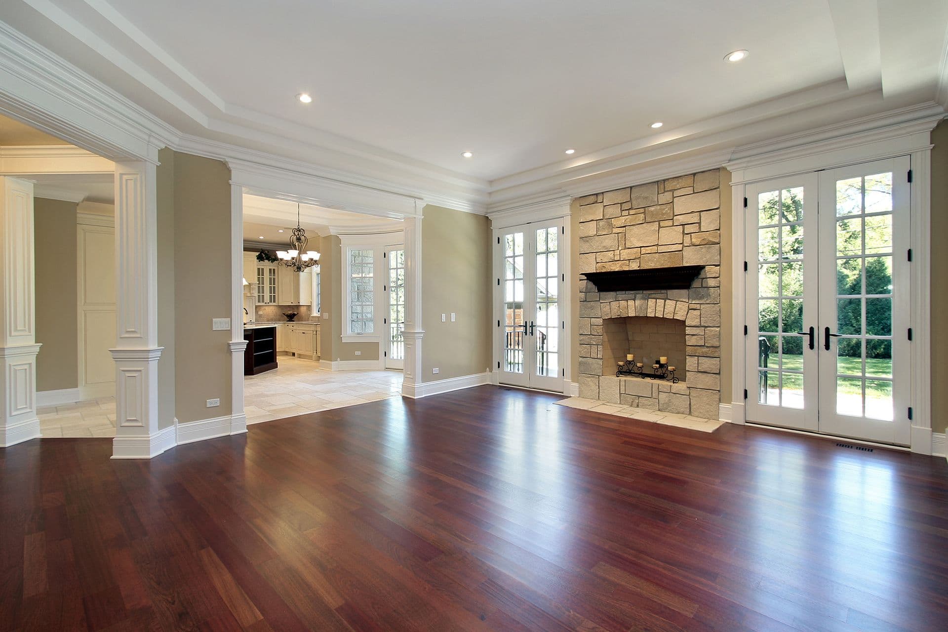 Spacious living room with stone fireplace, hardwood flooring, white coffered ceiling, French doors leading to patio, and open kitchen visible in background