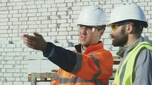 Two construction workers in hard hats and safety vests discuss a project in front of a brick wall, one pointing toward the work area